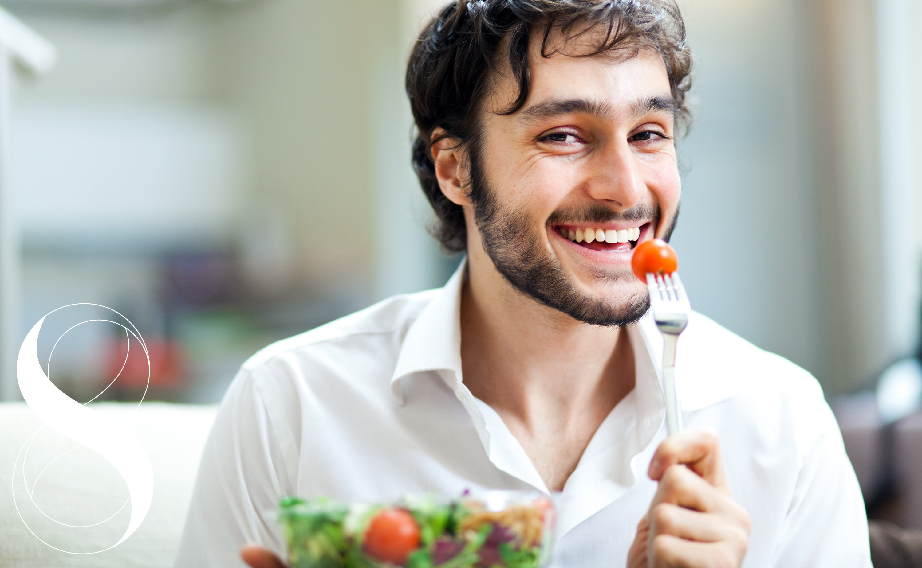 Ein lächelnder Mann mit dunklen, lockigen Haaren und Bart isst einen frischen Salat. Er hält eine Gabel mit einer Kirschtomate vor den Mund und blickt direkt in die Kamera. Ein Symbolbild von LIFE LIGHT für die Bedeutung eines stabilen Blutzuckers.