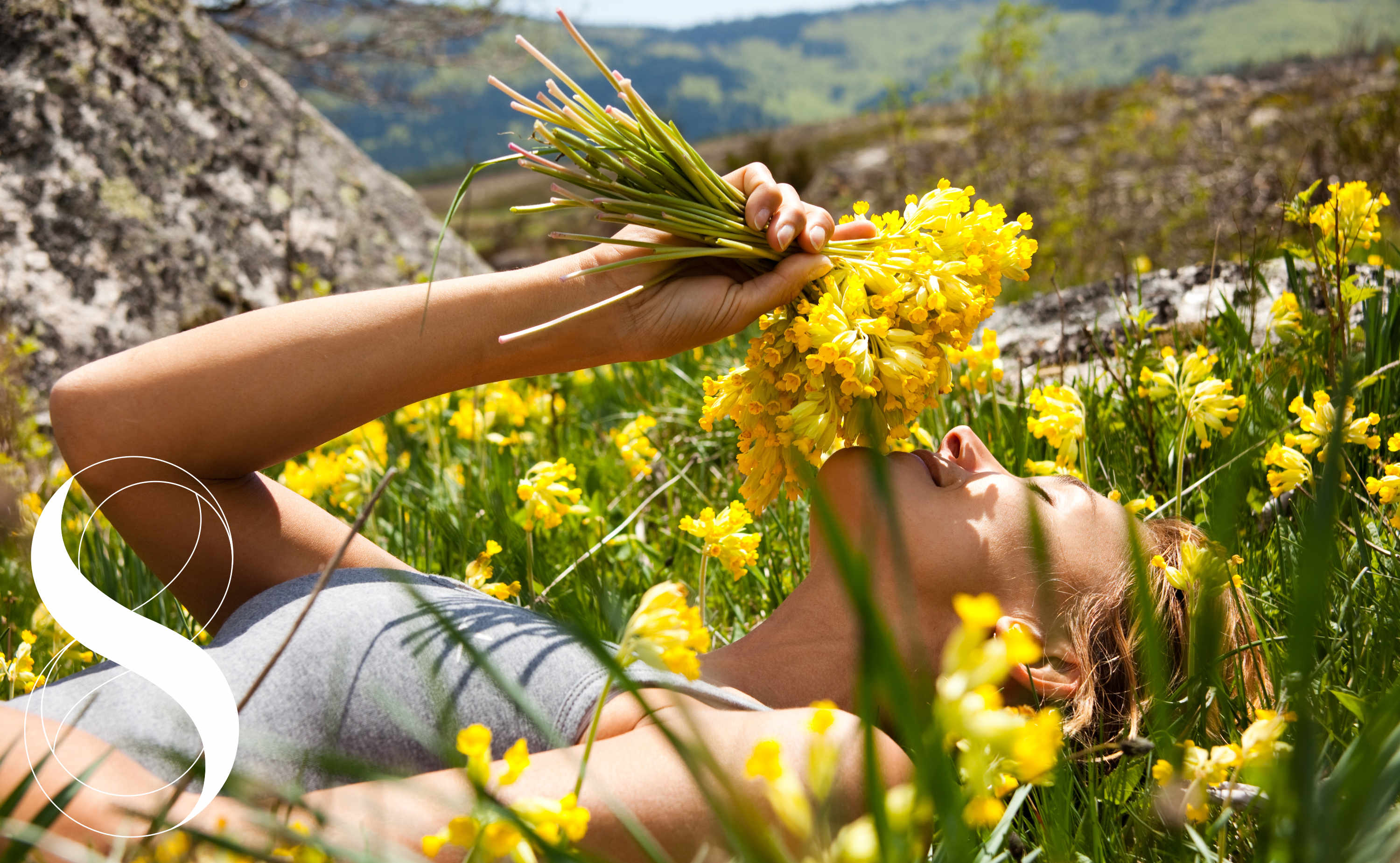 Eine Frau liegt entspannt in einer sonnigen Bergwiese voller gelber Schlüsselblumen und hält sich einen Strauß der Blumen nah ans Gesicht. Ein Symbolbild von LIFE LIGHT für Frühlingsgefühle statt Frühjahrsmüdigkeit.