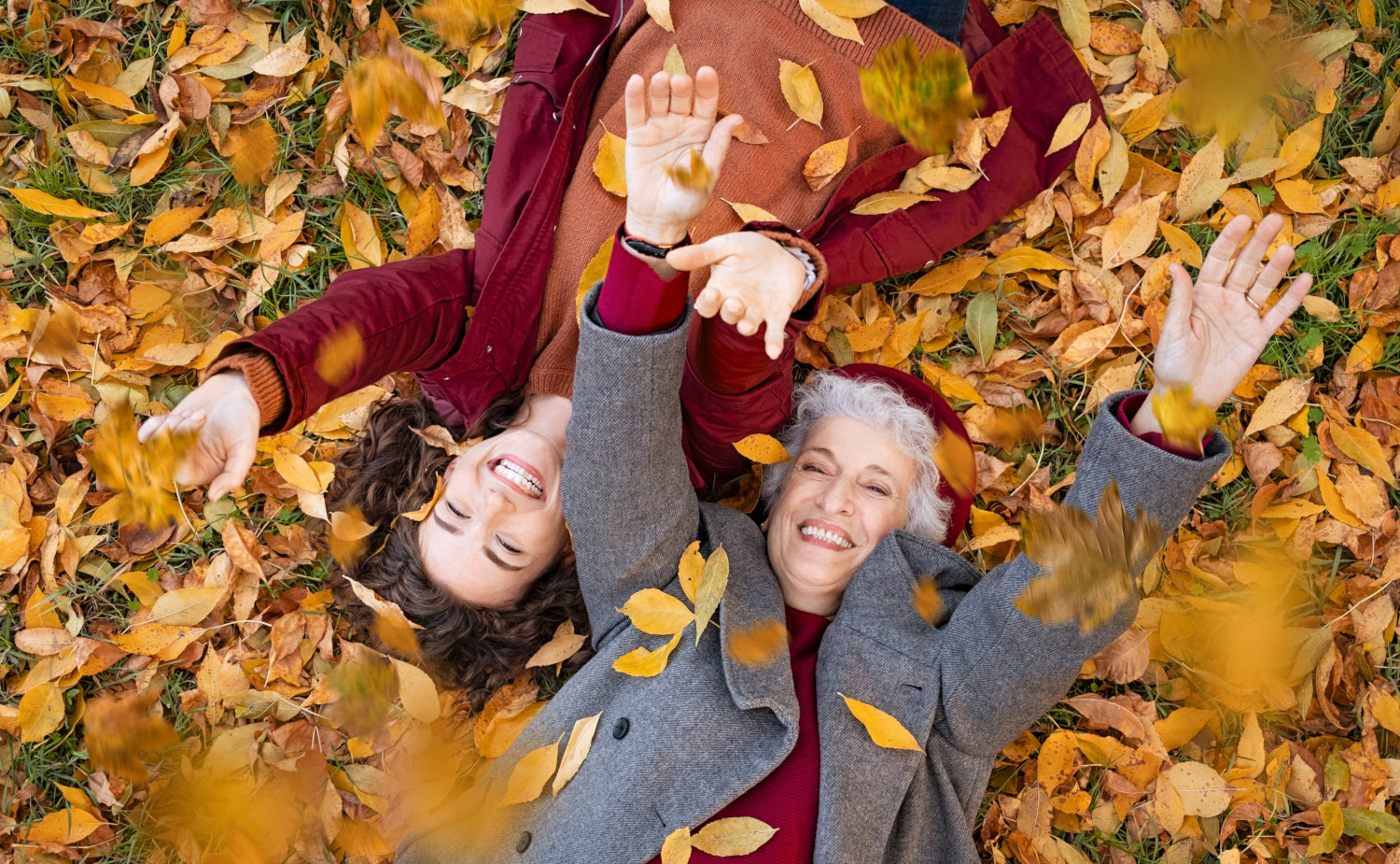 Zwei lächelnde Frauen liegen auf dem Rücken in einem Teppich aus herbstlichen Blättern, sie werfen Blätter in die Luft. Symbolfoto für starke Abwehrkräfte im Herbst.