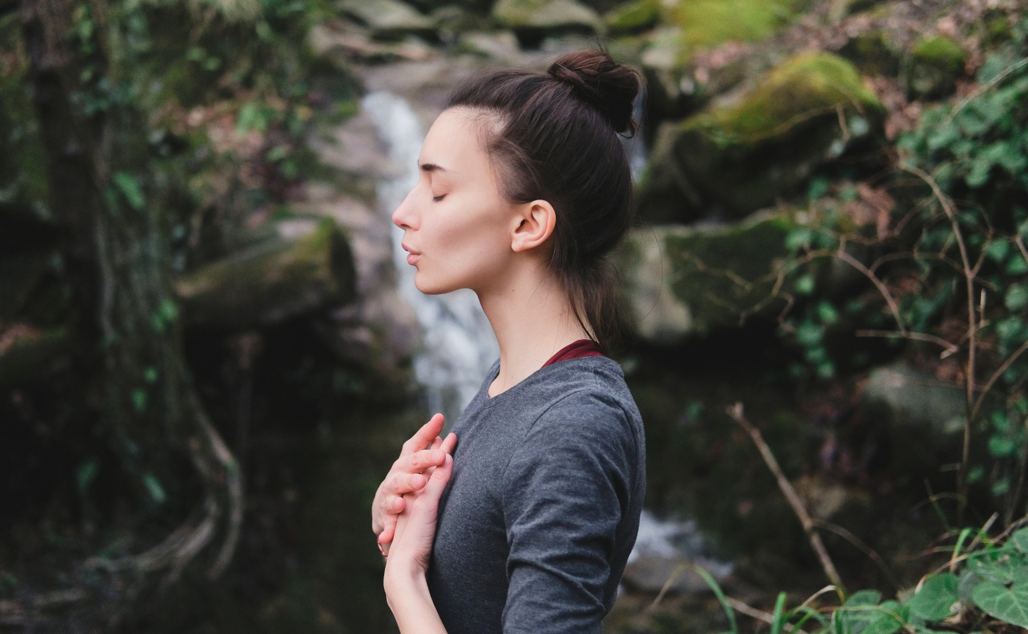 Eine Frau steht mit geschlossenen Augen und gefalteten Händen vor der Brust in der Natur, im Hintergrund ist ein kleiner Wasserfall. Symbolfoto für gesunde Lunge und frische Luft.