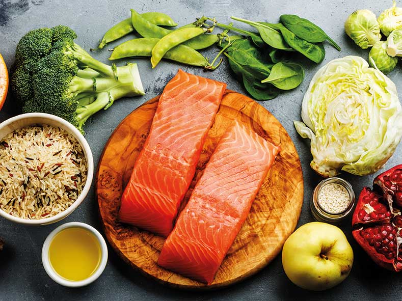 Lachs für ein gesundes Immunsystem Zwei Lachsfilets auf einem Holzbrett, umgeben von verschiedenen Gemüsesorten wie Brokkoli, Erbsenschoten, Spinat, Kohl und Granatapfel sowie Reis und Öl.