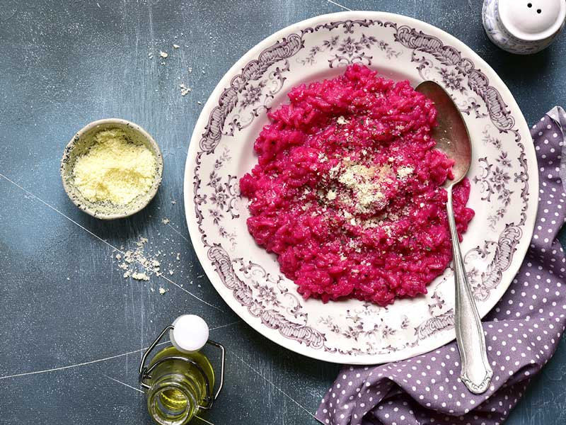 Rote Bete Risotto für ein gesundes Immunsystem Ein Teller mit leuchtend rotem Rote-Bete-Risotto, bestreut mit Käse, steht neben einer Schale mit geriebenem Käse und einer Flasche Öl.