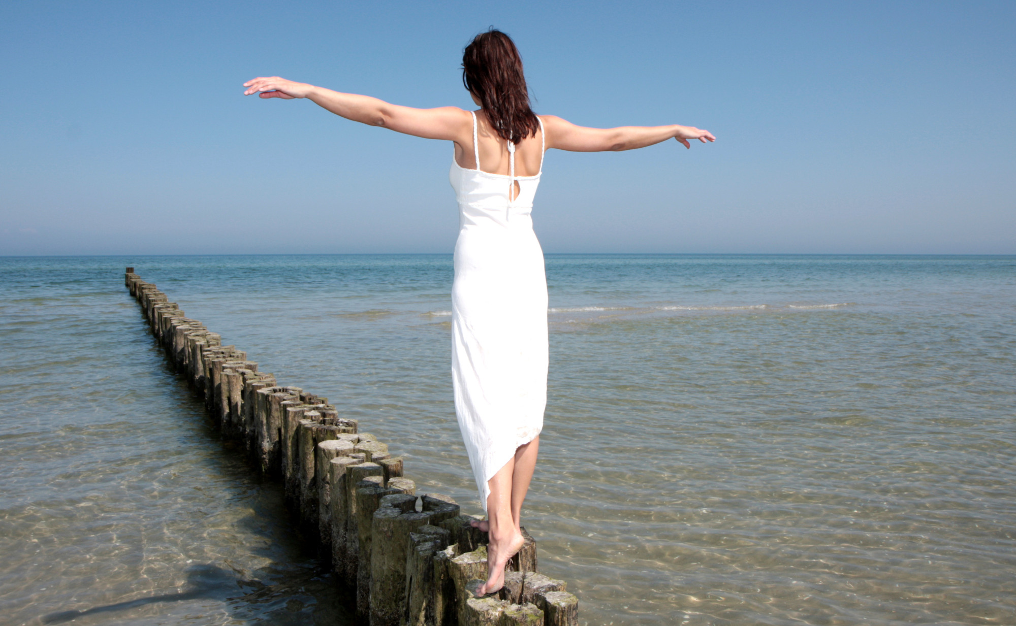 Eine Frau in einem weißen Kleid balanciert mit ausgebreiteten Armen auf einer Holzmole im Meer. Sie symbolisiert Balance - auch im Säure-Basen-Haushalt.