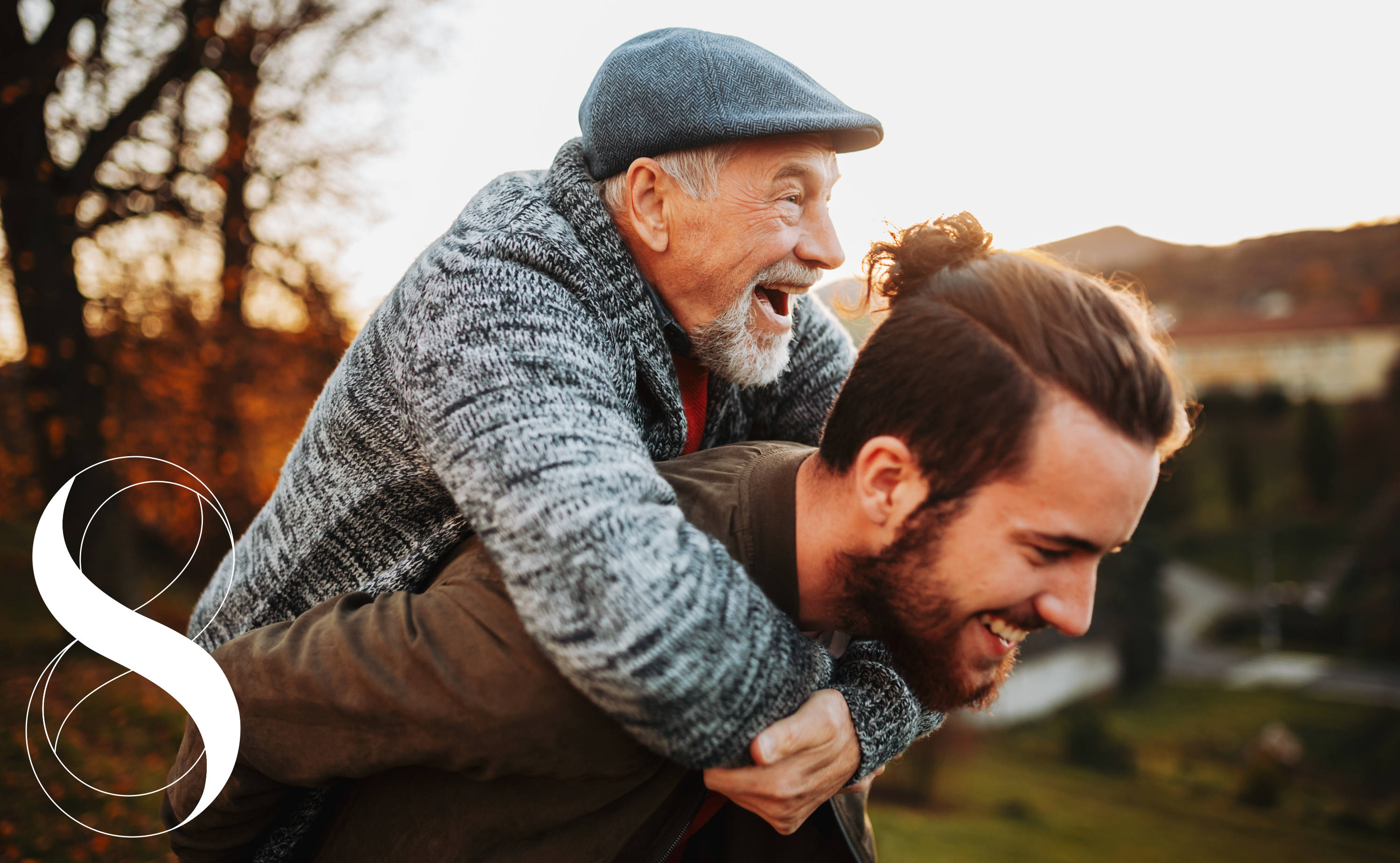 Ein fröhlicher älterer Mensch lässt sich lachend von einem jüngeren Menschen huckepack durch einen Park tragen. Ein Symbolbild für Lebensfreude, Agilität und Vitalität bis ins hohe Alter im Kontext von Zellgesundheit und Spermidin.