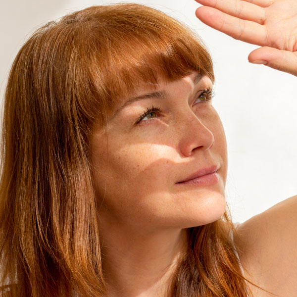 Rothaarige Frau hält schützend die Hand über die Stirn, um sich vor hellem Sonnenlicht zu schützen, das ihr Gesicht teilweise beleuchtet.