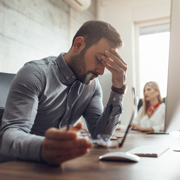 Ein Mann sitzt im Büro am Schreibtisch, hält eine Hand an seine Stirn und sieht gestresst oder erschöpft aus.