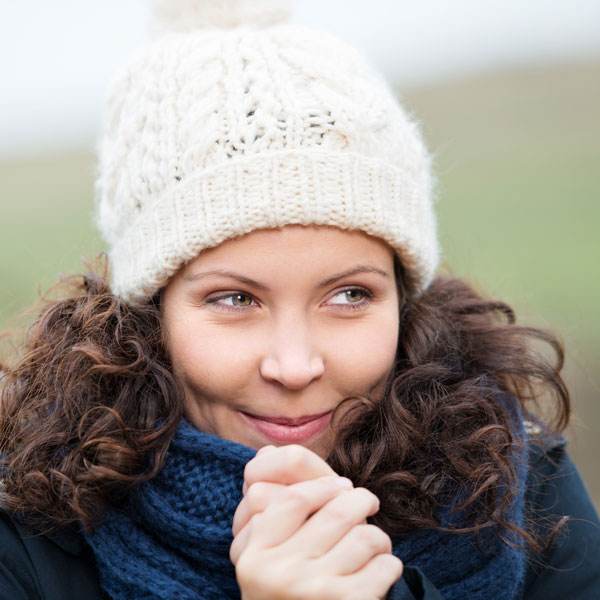 Eine junge Frau mit lockigem, braunem Haar, einer weißen Strickmütze und einem blauen Schal, die leicht fröstelt und sich die Hände reibt.