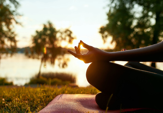 Ein Mensch meditiert im Sonnenuntergang am See, mit Hand in Mudra-Stellung auf einer Yogamatte. Ein Symbolfoto von LIFE LIGHT für Energie und gesunde Zellen in Balance.