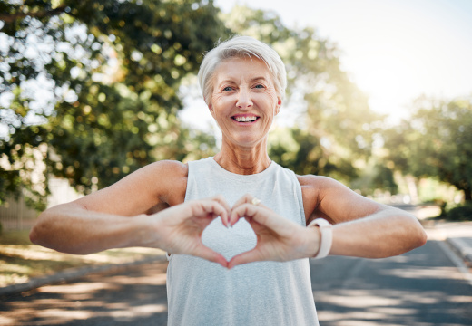 Eine lächelnde Frau mit kurzen, grauen Haaren in Sportkleidung, ihre Hände formen ein Herz. Im Hintergrund sind Bäume und die strahlende Sonne. Ein Symbolfoto von LIFE LIGHT zur Herzgesundheit an heißen Tagen.