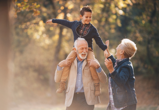 Großeltern und Kind im Herbstwald: Ein Kind sitzt auf den Schultern des Großvaters, während sich die Großmutter daneben zum Kind hochstreckt und lacht. Ein Symbolbild von LIFE LIGHT für Zellschutz und gesundes Älterwerden.